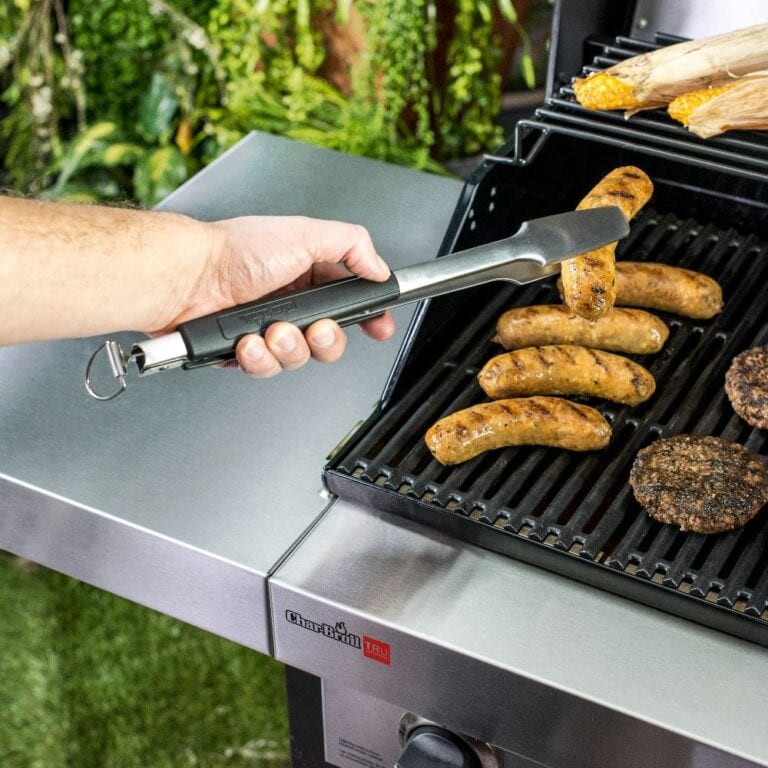 Metal kitchen tongs held by hand while grilling burgers and sausages outdoors.